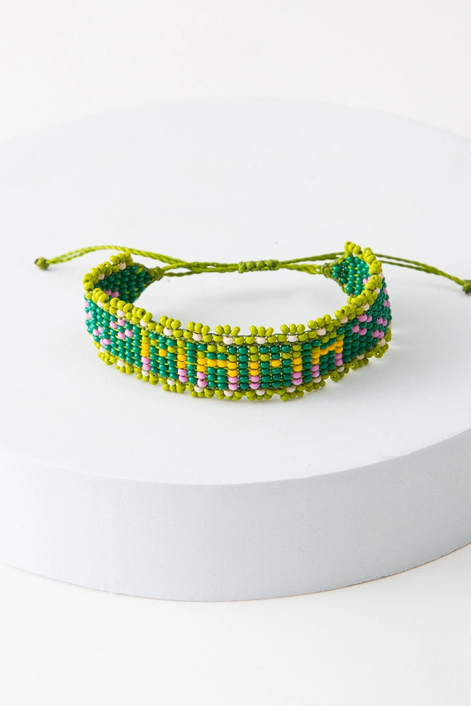 The Mama Beaded Bracelet is an adjustable thread bracelet featuring a band of green glass beads. Within the band, the word "Mama" is formed with lime, yellow, and pink glass beads. A row of lime glass beads forms a border around the bracelet. A lime thread closure at the back of the bracelet can be adjusted by pulling on both ends of the thread.