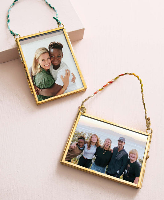 The two frames included in the Memento Frame Ornaments set lay on a peach background. Each of the glass frames is outlined in shining brass. Both frames are rectangular, but one is oriented vertically and the other horizontally. On one edge of each frame, a mechanism enables the brass frame to open so that a photo may be placed between the glass and the frame. A twisted cord made of multicolor fabric is connected to the top corners of each frame, allowing them to be hung.