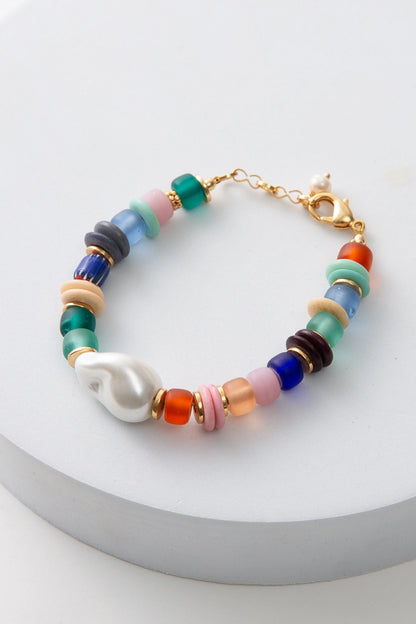 The Menagerie Bracelet is a single strand bracelet composed of a rainbow of glass beads. The beads include shades of red, pink, orange, blue, and green. Disc-shaped brass and bone beads are interspersed with the glass beads. A glass bead in the shape of a freshwater pearl sits at the front of the bracelet. The bracelet has a gold clasp.