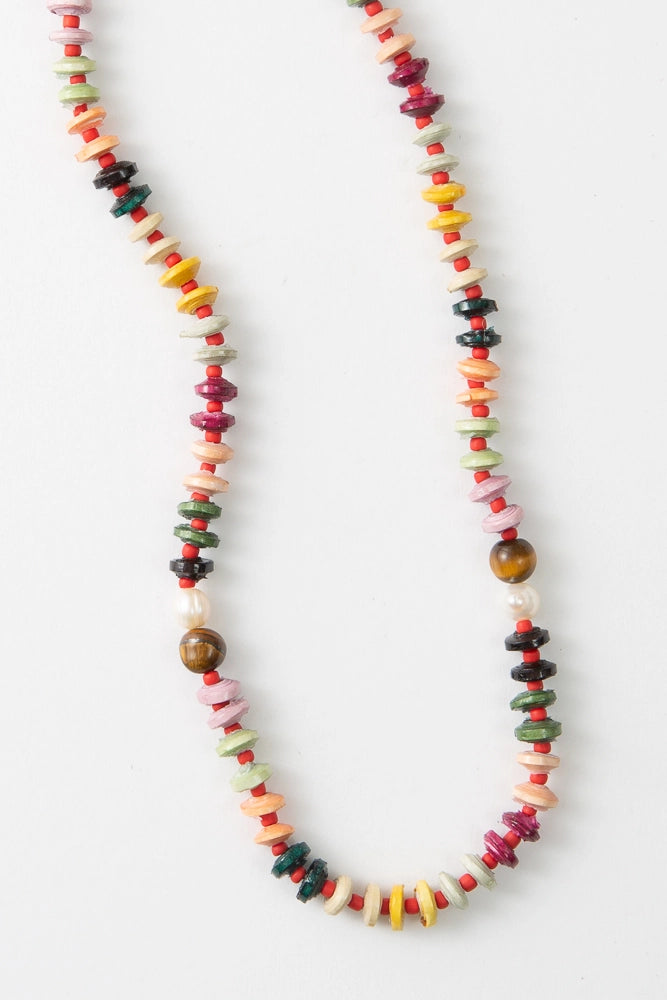 The Mihingo Necklace is a short beaded rope necklace. It is composed of many different colored disc-shaped paper beads. The beads are pink, mint, dark green, yellow, cream, and orange. The disc-shaped beads alternate with small red glass beads. Near the middle of the necklace there are accents of pearl and tiger's eye beads. 