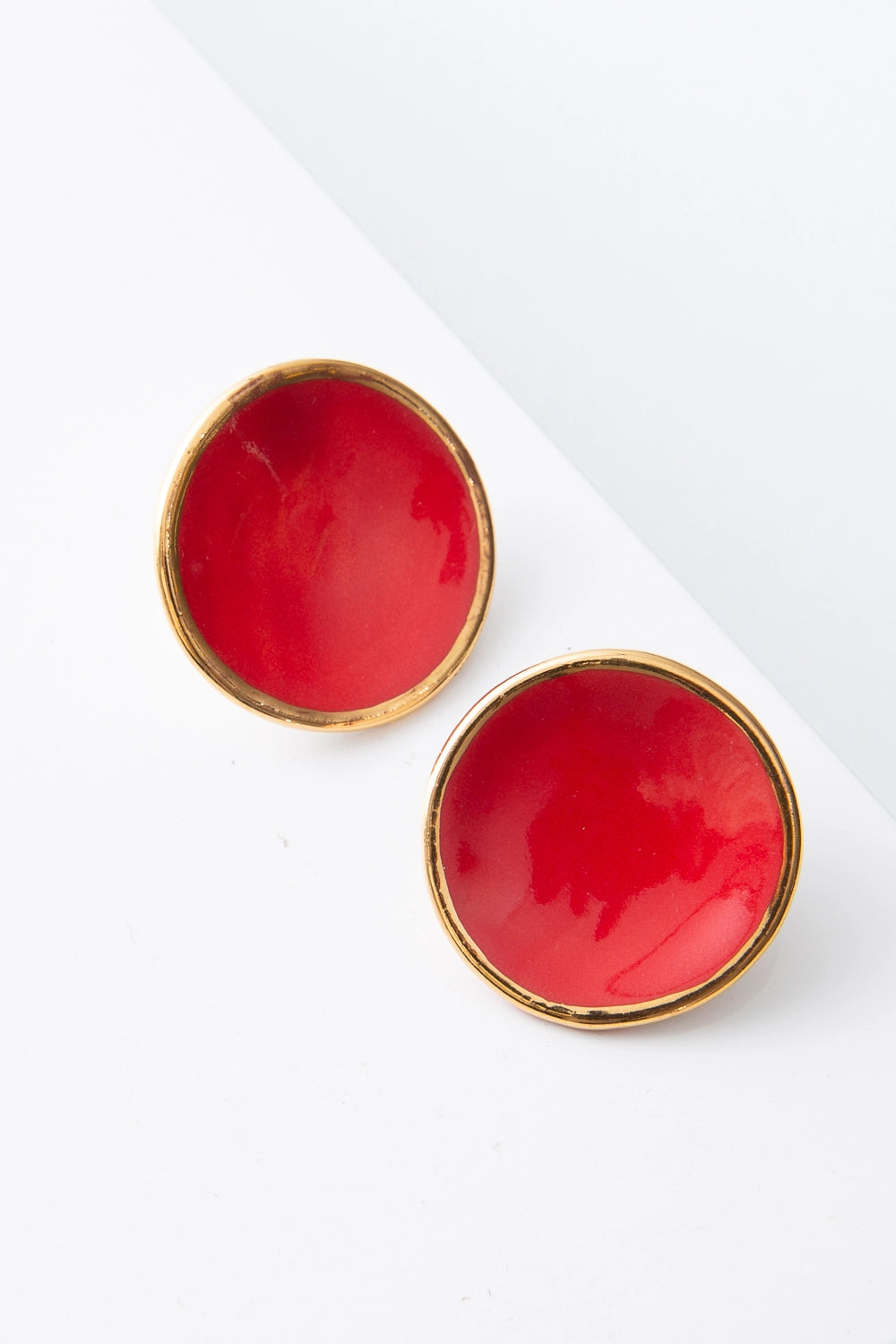 The Mini Halo Earrings are flat, disc-shaped post earrings composed of bright red porcelain. The small discs are outlined in a ring of shining gold. The porcelain has natural variations and subtle markings, giving it a handmade look.