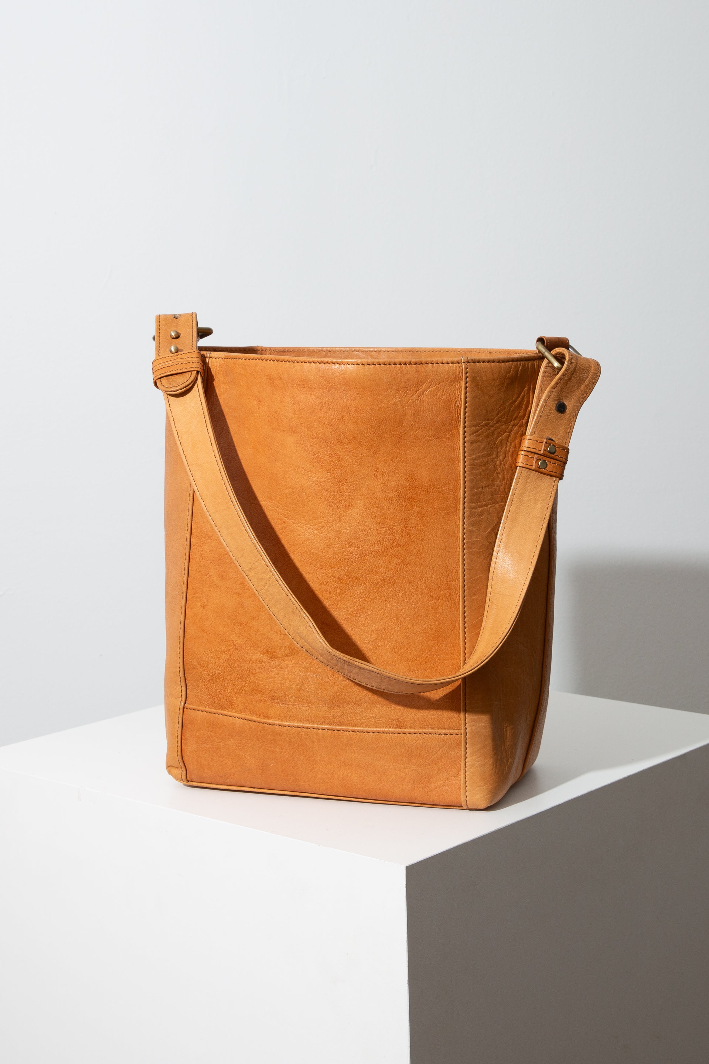 This is a rectangular leather tote bag that is taller than it is wide. It is composed of smooth, caramel-colored leather. The top of the bag is open. A matching, adjustable leather strap is attached to the two top corners of the bag.