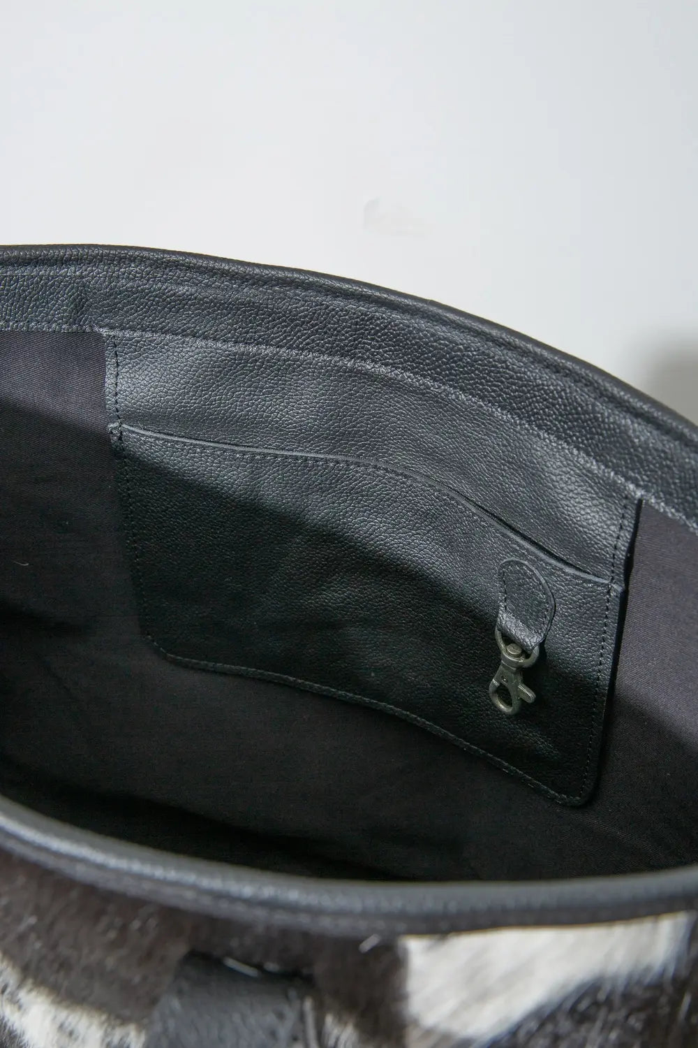 The interior of the Modern Leather Cowhide Tote is shown. It has a black fabric lining and an interior leather pocket stitched into the back wall of the bag. The leather pocket has a clip attached where a keyring or other item can be attached.