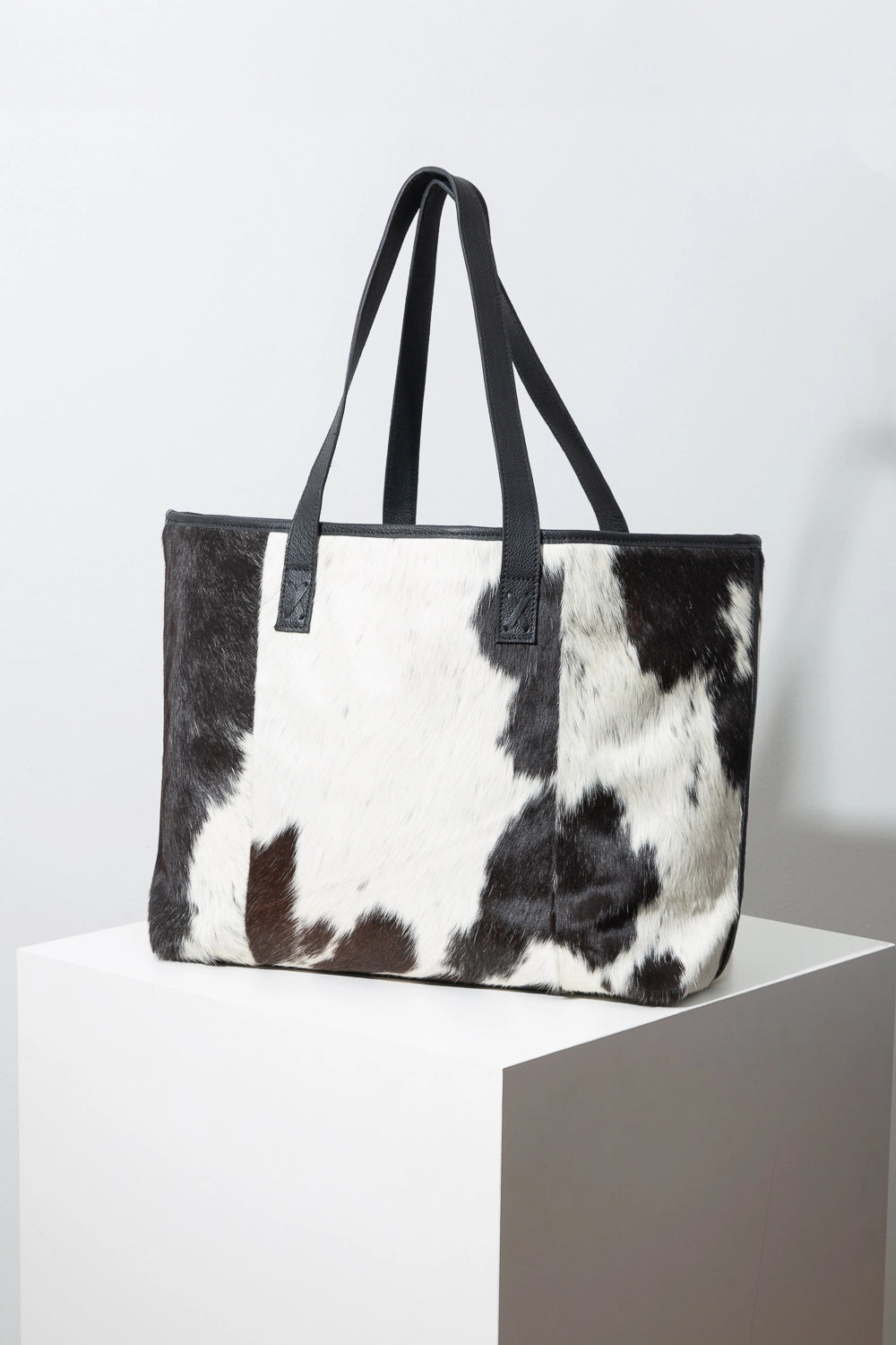 This is a large leather tote that is wider than it is tall. The bag is composed of black and white patterned cowhide. There are two black leather shoulder straps at the top of the bag. The tote is open at the top.