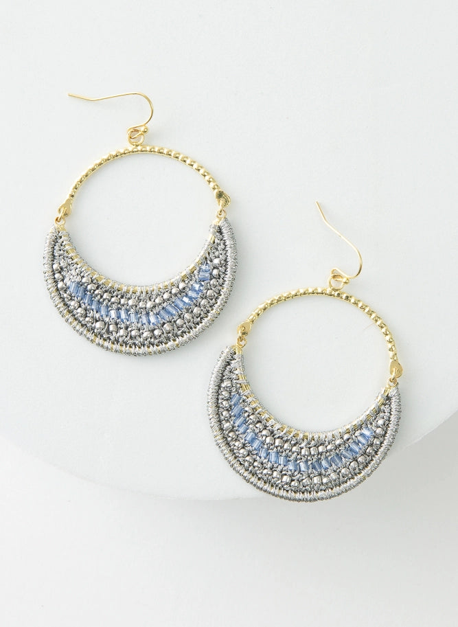 The Moonlight Earrings are hoop earrings. The top half of the hoop is gold metal. The bottom half of the hoop is filled with a crescent of silver thread and light blue glass beads.