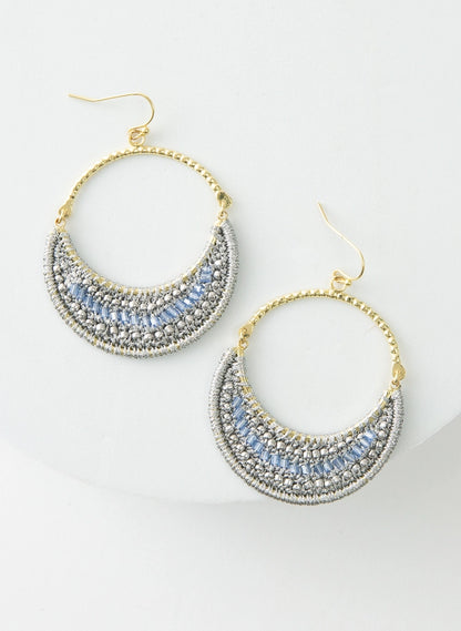 The Moonlight Earrings are hoop earrings. The top half of the hoop is gold metal. The bottom half of the hoop is filled with a crescent of silver thread and light blue glass beads.