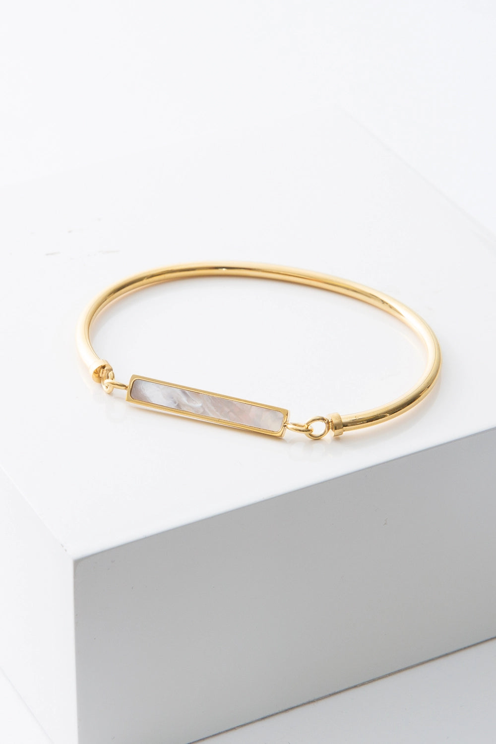 This is a thin cuff composed of one solid piece of gold-plated stainless steel that connects on either side to a narrow rectangular pendant that is suspended at the front of the cuff. The pendant is composed of shining mother-of-pearl outlined in gleaming gold.