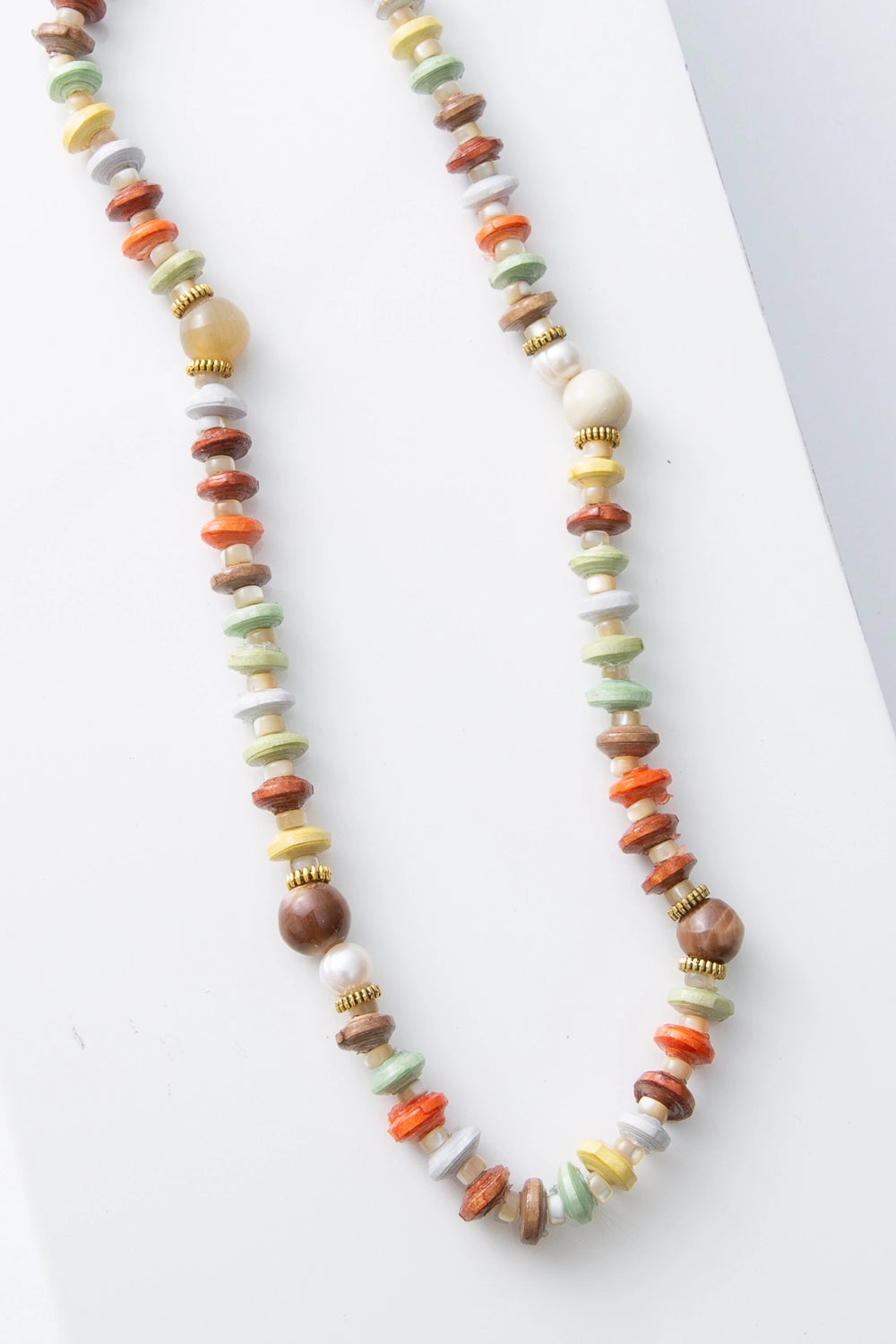 This is a single-strand rope necklace that features a variety of bead colors, shapes, and materials. Disc-shape paper beads in shades of brown, orange, mint, yellow, and cream alternate with small ox horn beads. Every few inches, the necklace is dotted with round pearl and ox horn beads.