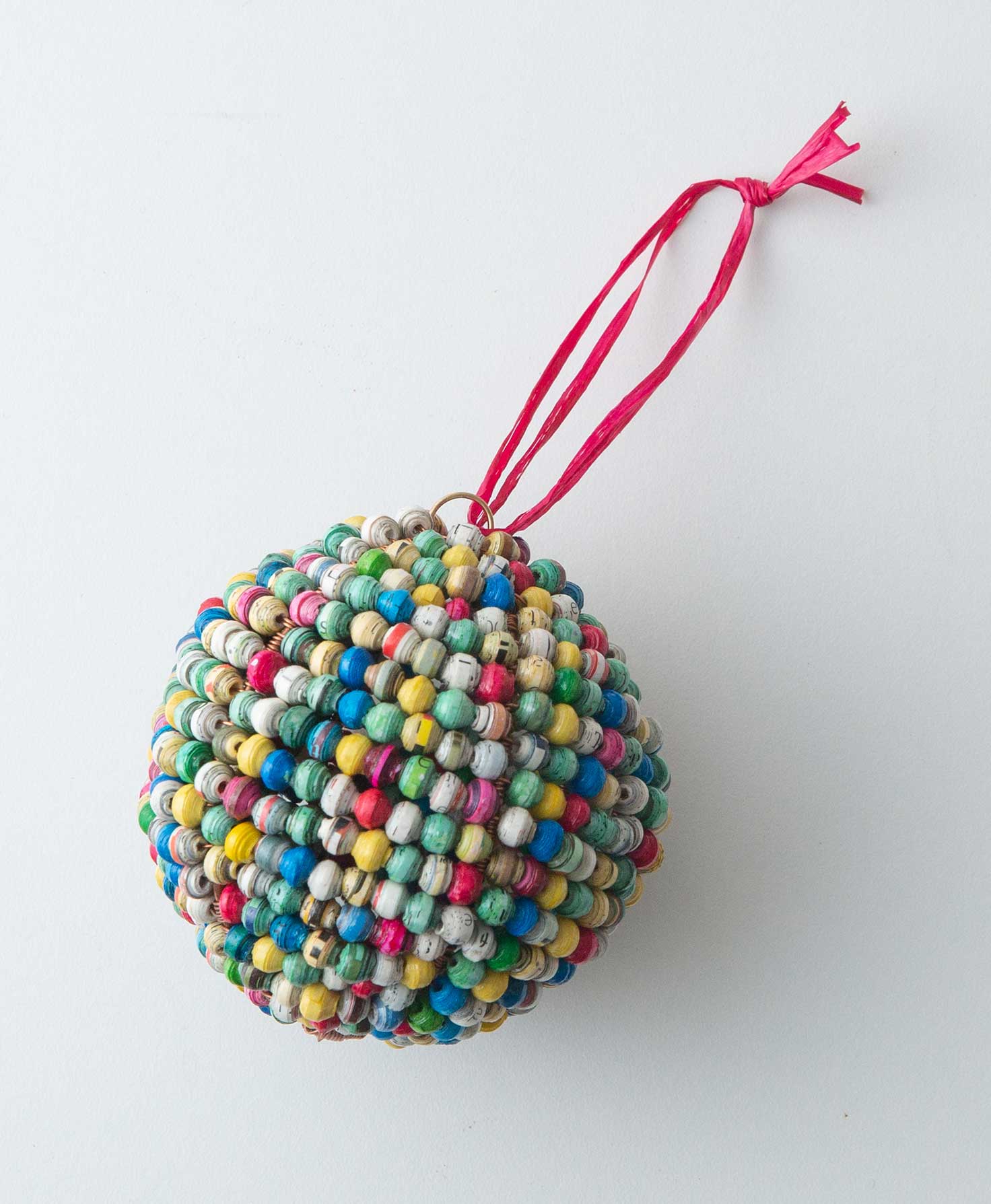 The Paper Sphere Ornament is a sphere shaped ornament composed entirely of small handrolled paper beads. The beads are every color of the rainbow. At the top of the ornament there is a red raffia loop that allows it to be hung.