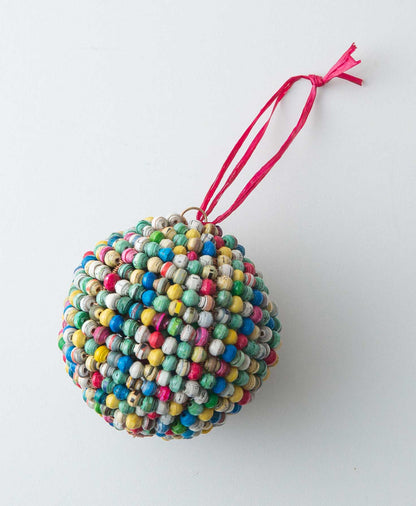 The Paper Sphere Ornament is a sphere shaped ornament composed entirely of small handrolled paper beads. The beads are every color of the rainbow. At the top of the ornament there is a red raffia loop that allows it to be hung.