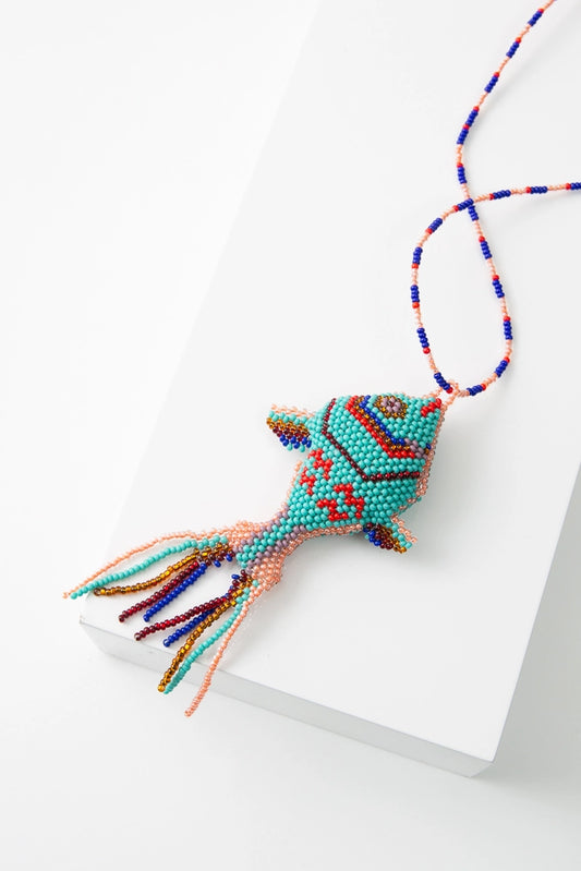 The Pescado Bag Charm is a three-dimensional beaded fish charm. The base of the fish is turquoise glass beads, and the fish's features are depicted using red, blue, pink, and purple beads. There is a beaded fringe at the fish's tail. The charm is attached to a beaded loop that allows it to be attached to a bag or other item.