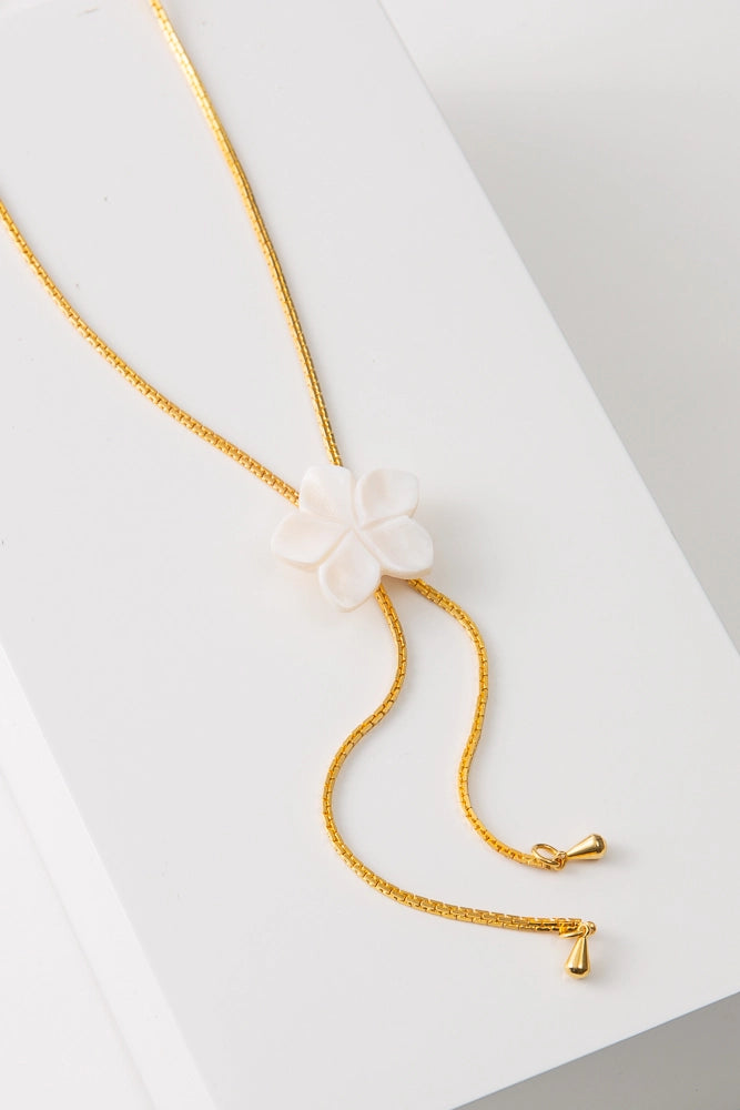 The Plumeria Lariat Necklace has a gold chain that loops around the back of the neck and is attached at the front with a white flower clasp. The flower is composed of mother of pearl and has a minimalist design. The flower can slide up or down to create different looks.