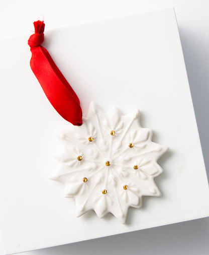 The Porcelain Snowflake Ornament lays against a white background. It is a flat snowflake-shaped ornament made of shiny white porcelain. Gold dots accent the different points of the snowflake. A silky red ribbon attached to the top allows the ornament to be hung.