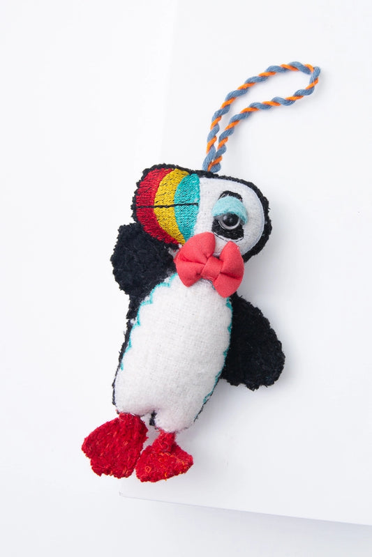 The Puffy Puffin ornament is a three-dimensional wool ornament in the shape of a puffin. The body is black and white, the feet are red, and the bill is red, yellow, and blue. The puffin has a shiny black eye and a red bowtie. There is a twisted fabric cord at the top of the ornament where it can be hung.