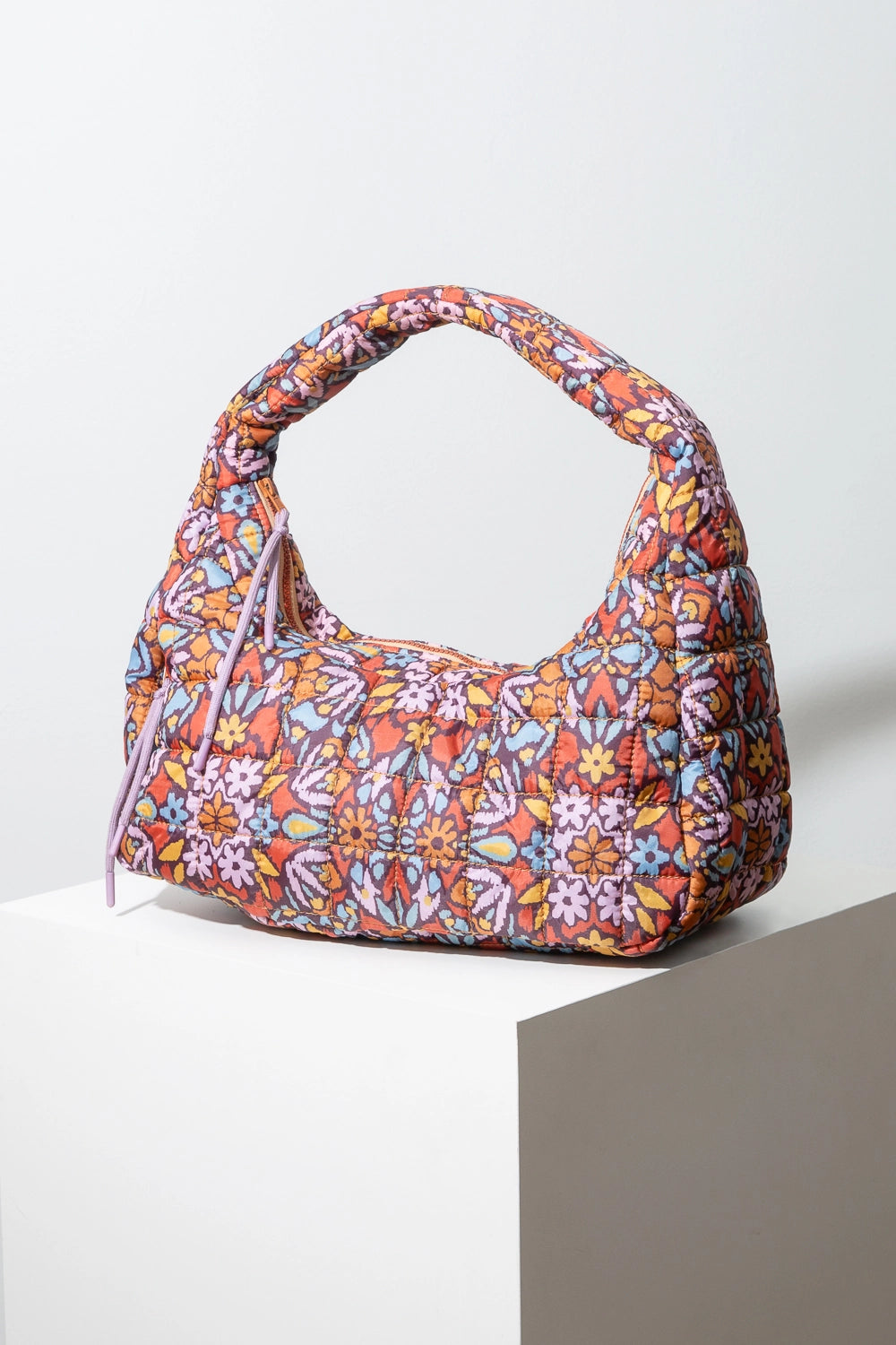 This is a soft fabric handbag with a quilted texture. It is wider than it is tall. The fabric has a bright, mosaic-like floral print. It has a red-orange background with orange, blue, yellow, and lavender floral designs. A gold zipper stretches across the top of the bag and there is a zipper pocket on the front of the bag. Both zippers have lavender string tie pulls. The handle of the bag is composed of the same soft floral print quilted fabric.