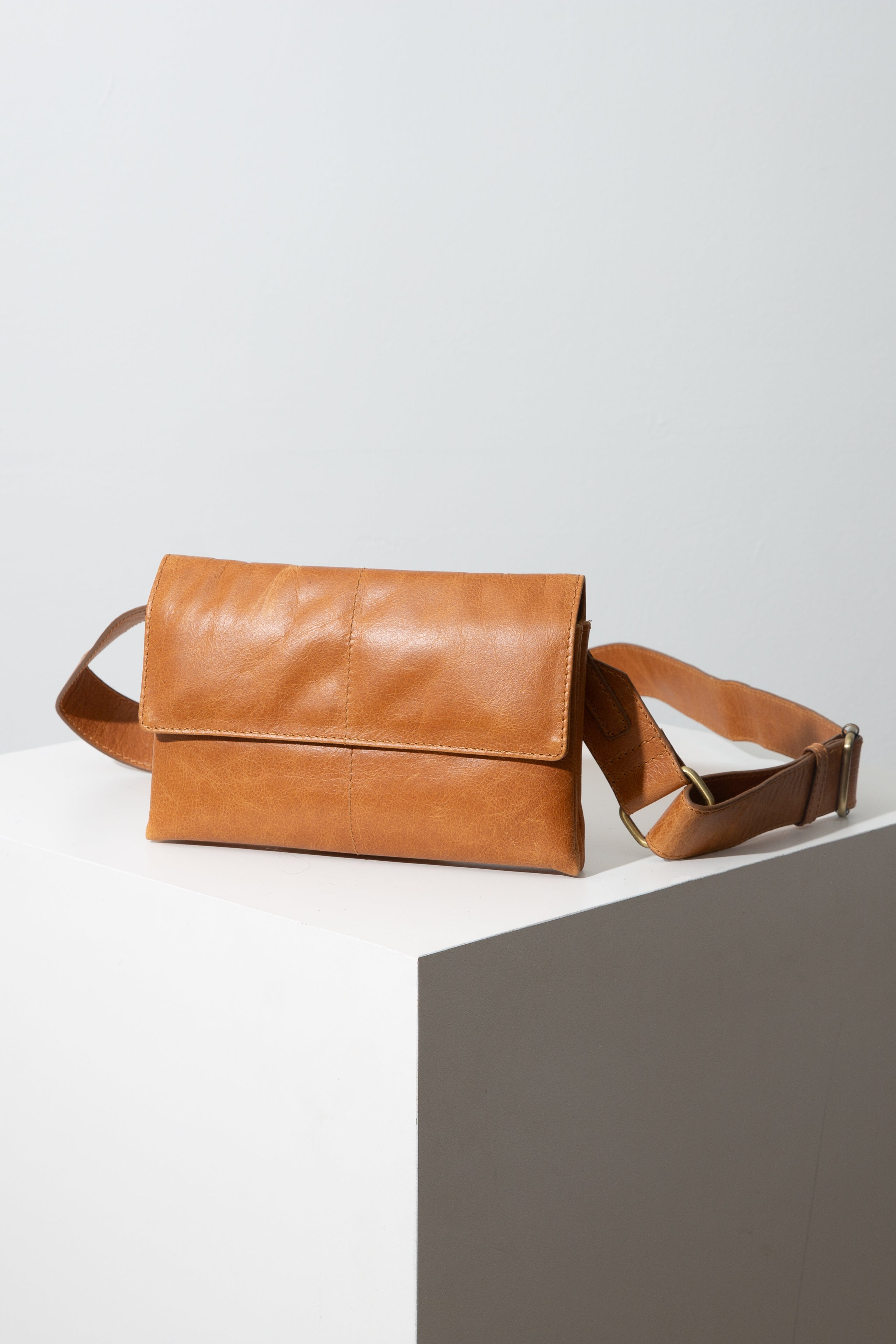This is a rectangular crossbody bag that is wider than it is tall. It is composed of smooth, caramel-colored leather. It has a flap that folds over and covers half of the front of the bag, where it is clasped in place with a magnetic closure. The leather is stitched in a way that resembles a quilt, with lines of stitches running around the perimeter of the bag and down the center.  A long adjustable leather strap is attached to the top corners of the bag.
