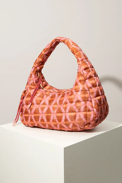 The Quilted Zipper Tote is a soft, oversized handbag composed of quilted fabric. The fabric features an orange and pink tie-die style geometric print. the bag has a crescent shape with a short handle. There is a zipper closure finished with peach cords. There is a vertical zipper pocket stitched into the front of the bag.