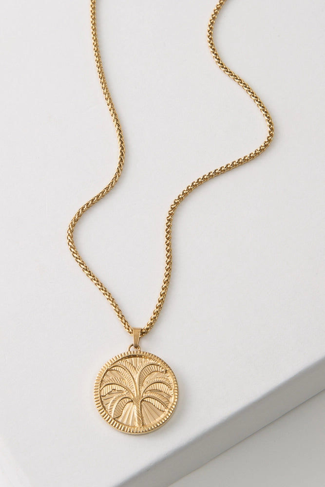 The Recharge Medallion is a gold pendant necklace. A round, gold pendant hangs from a gold chain. The pendant is etched with the image of a palm tree rising above a sand dune. The border of the pendant is etched with tiny lines, giving it a coin-like look.