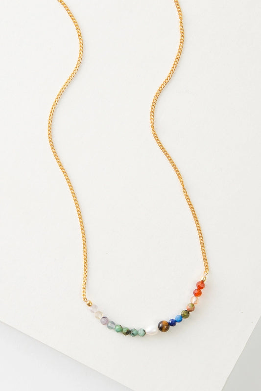 The Refraction Necklace is a short gold chain necklace. There is a row of small beads at the base in all colors of the rainbow. The beads are composed of crystal, semi-precious stones, brass, and pearl.