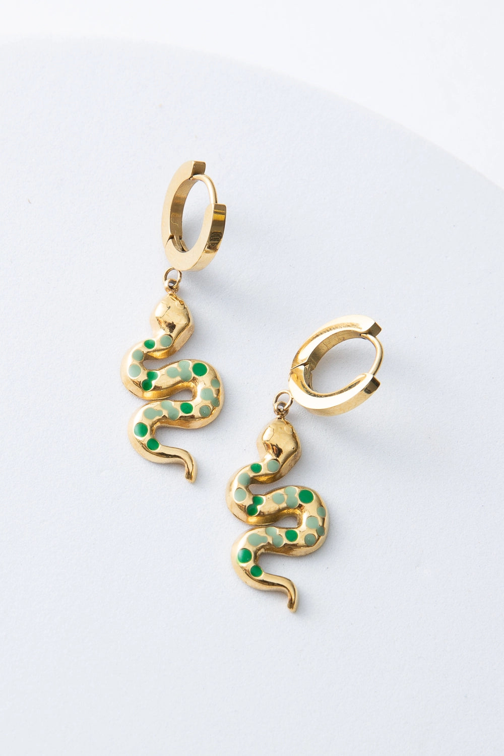 These are huggie-style earrings with a gold hoop that fits snuggly around the ear lobe. Hanging from the hoop is a gold snake charm dotted with green and light green enamel speckles. The body of the snake curls back and forth, forming an S-shape.