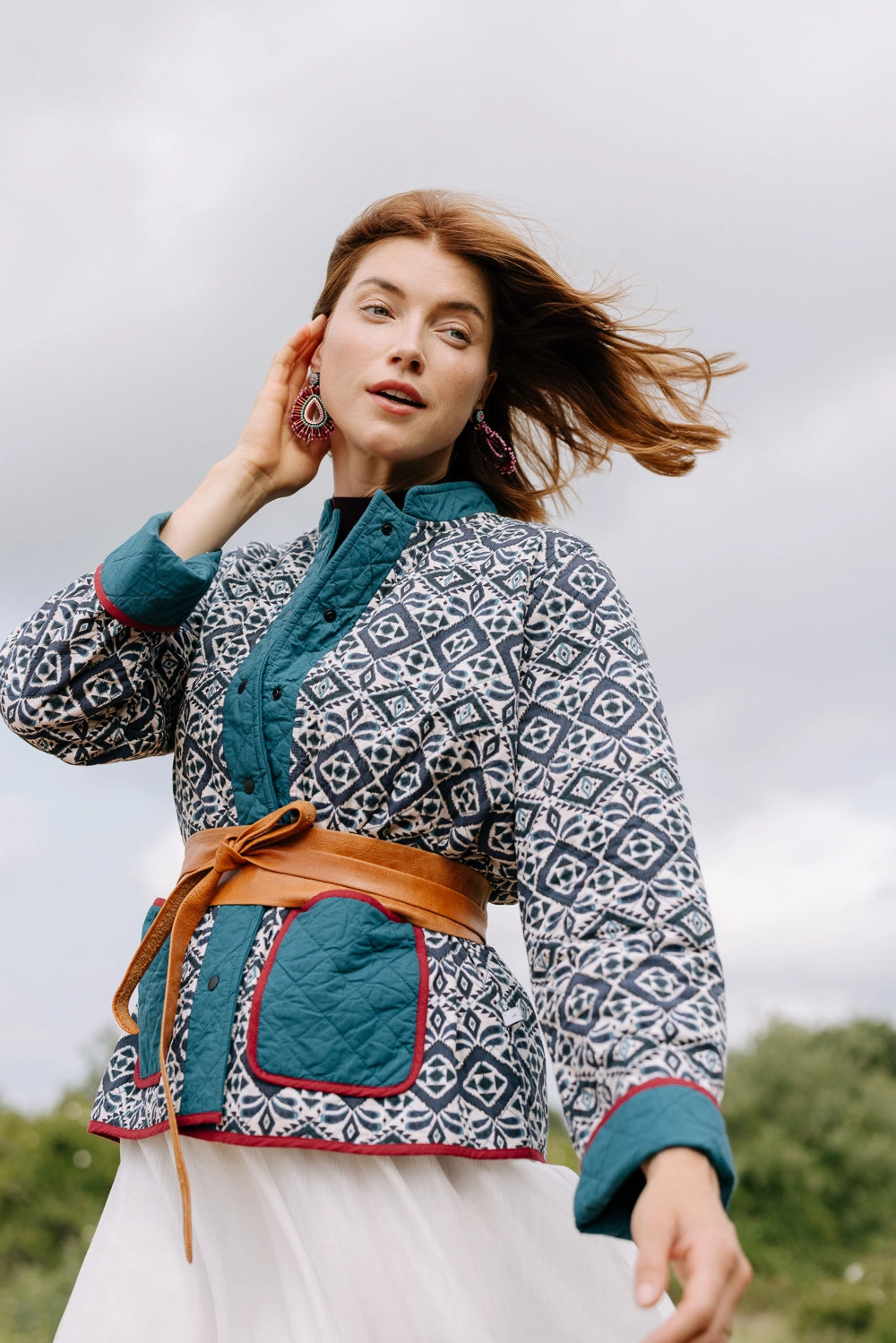 This jacket has a quilted texture and is composed of 100% cotton. The bulk of the jacket is a mosaic-style navy and white print fabric, and the collar, cuffs, button closure, and pockets are teal fabric with a red cord border. There are two large pockets that are open at the top. The jacket has black snap buttons along the front as a closure. The model wears the jacket belted at the waist with a leather wrap-around belt.