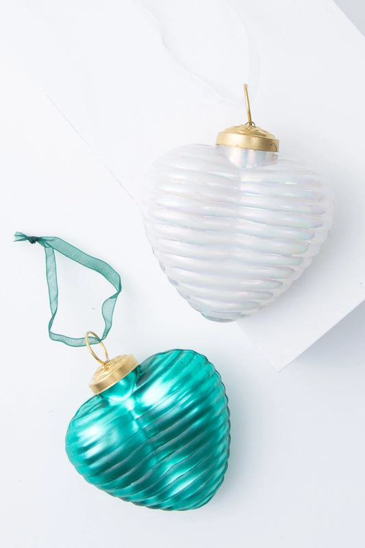 The two ornaments included in the Ribbed Glass Heart Ornament set are pictured. They are each three-dimensional heart ornaments made of glass with a horizontal ribbed pattern all around them. One ornament is teal and one is white. Both have a metallic shine. There is a brass hook at the top of each ornament with a matching ribbon loop attached.