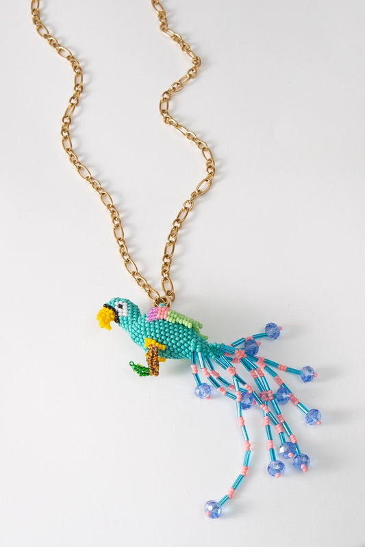 The Rico Necklace is a gold chain necklace with a beaded bird pendant. The three-dimensional bird is composed of brightly colored glass beads. The base of the bird is turquoise, the beak is yellow, and the wings are green and pink. A long beaded fringe tail extends from the bird's tail feathers in shades of blue and pink.