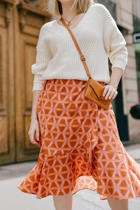 The Ruffle Wrap Skirt in Munni Sunset is a mid-length wrap skirt composed of a flowy rayon-cotton blend fabric. The fabric features an orange and pink tie-die style geometric print. The skirt hits just below the knee and has a ruffled hem.