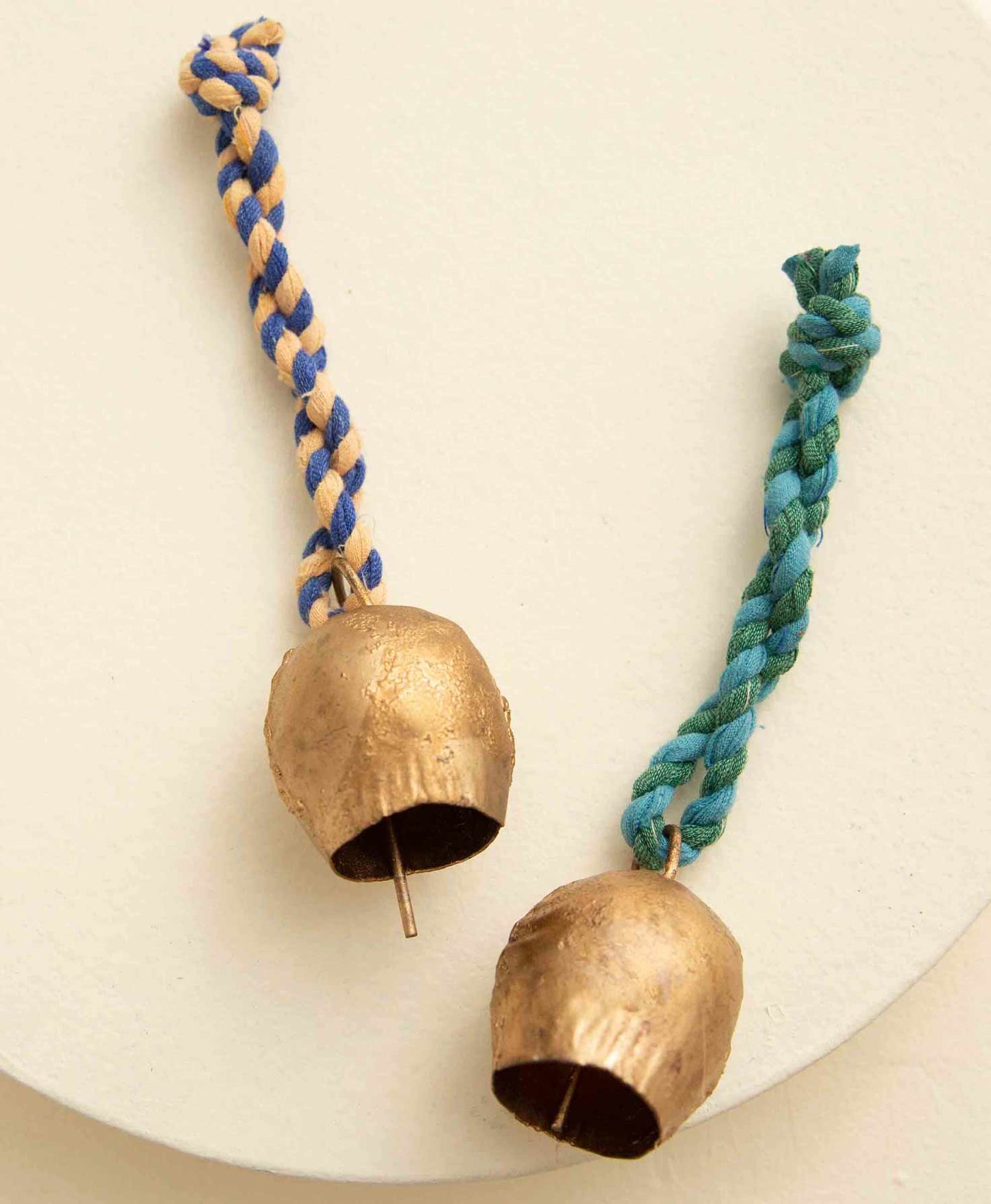 The two ornaments included in the Rustic Bell Ornament set have the same look with different color palettes. Both feature a small, rustic brass bell with a handmade look. Attached to each bell is a twisted fabric loop that can be used to hang the ornament. One ornament has a peach and blue loop, while the other has a green and aqua loop.