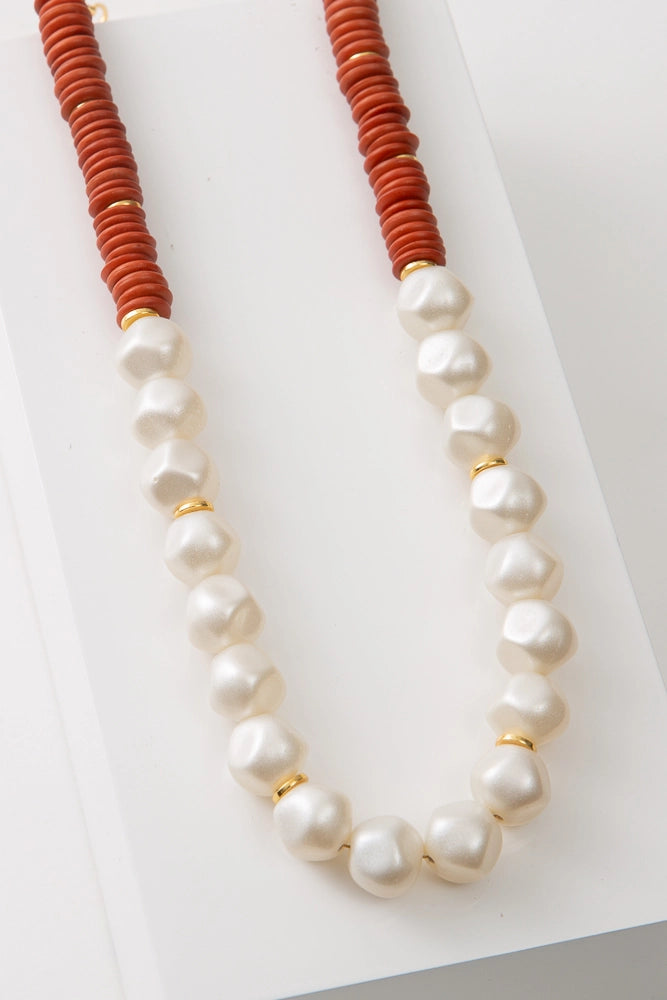 The Sahil Necklace is a single strand necklace. The top half of the necklace is strung with rust-colored disc-shaped beads. The bottom half is strung with iridescent white beads resembling roughly polished pearls. Brass discs are strung at regular intervals between the beads.