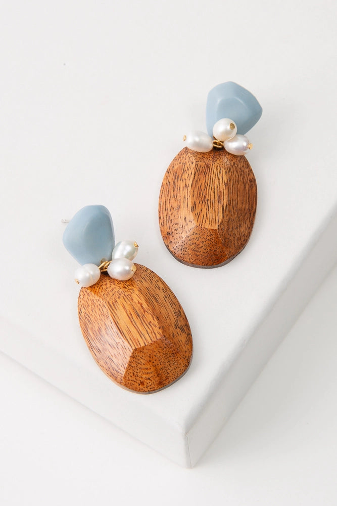 The Salinas Earrings feature a light blue, faceted tagua seed piece attached to a silver ear post. Below this hangs a faceted wood oval cut to resemble a gemstone. Three natural pearl charms are attached between the tagua and wood pieces.