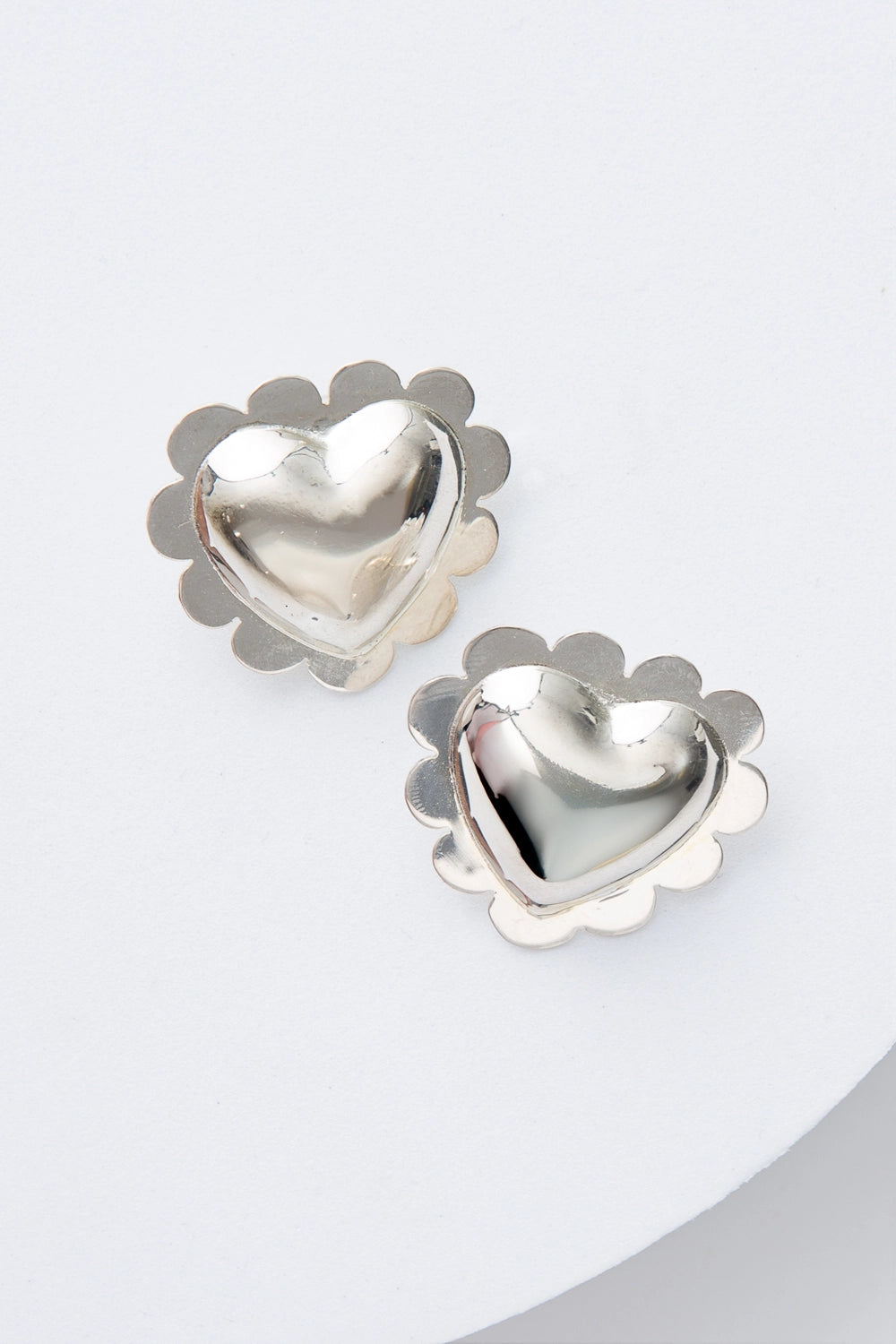 The Scalloped Heart Earrings are shiny silver earrings in the shape of puffy, three-dimensional hearts. There is a silver scalloped border around the outside of the heart shapes. They are stud earrings. 