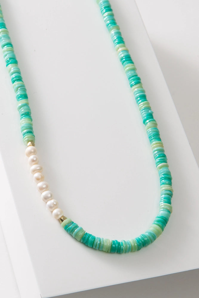 The Sea Pearl Necklace is a single stranded necklace composed of tiny disc-shaped shell beads in varying shades of turquoise. At one section of the necklace there is a row of eight round pearls with a faceted gold bead on each side.