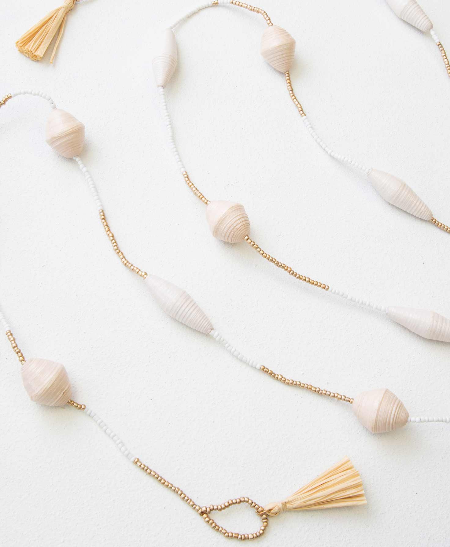 The Serenity Paper Bead Garland is composed of a single strand of beads that alternates sections of gold and white beads. Every few inches there is a large hand-rolled paper bead in either white or cream. There are natural sisal tassels at each end of the garland, along with a loop that can be used to attach the garland.