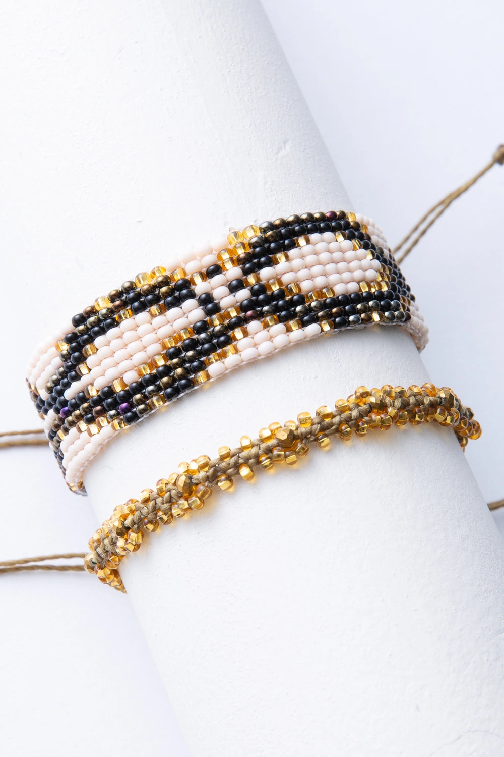 The two bracelets included in the Serpiente Bracelet Set are shown together. Both are composed of glass beads and have a pull-string that can be adjusted for size. One bracelet is composed of cream, black, and shiny gold glass beads woven to form a mosaic pattern. The other bracelet is thinner and is composed of a single cord embellished with gold and brass beads.