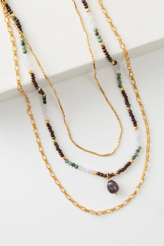 The Sharma Necklace is a three-stranded necklace. The shortest strand is composed of small gold beads. The middle strand is composed of semi-precious stone, quartz, and wood beads with a dark pearl charm. The longest strand is a gold metal chain.