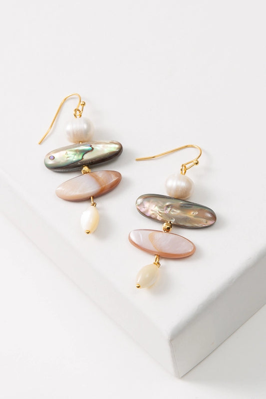 The Shell Trove Earrings are composed of gold ear hooks with pearl and shell pieces hanging down vertically, giving them a stacked look. At the top of the stack is a round pearl. Below this is an iridescent black shell piece, followed by a slightly narrower iridescent peach shell piece. At the bottom of the stack is an oval-shaped pearl bead.