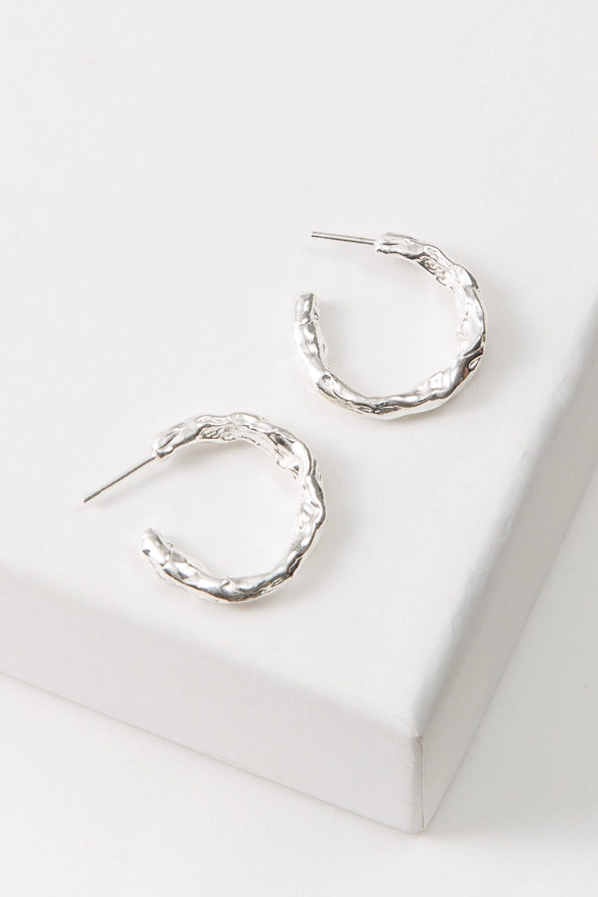 The Molten Metal Hoops are midi-sized hoops composed entirely of shiny silver metal. The metal has a rough, irregular shape that gives the earrings an organic feel. The hoops are open at the back.