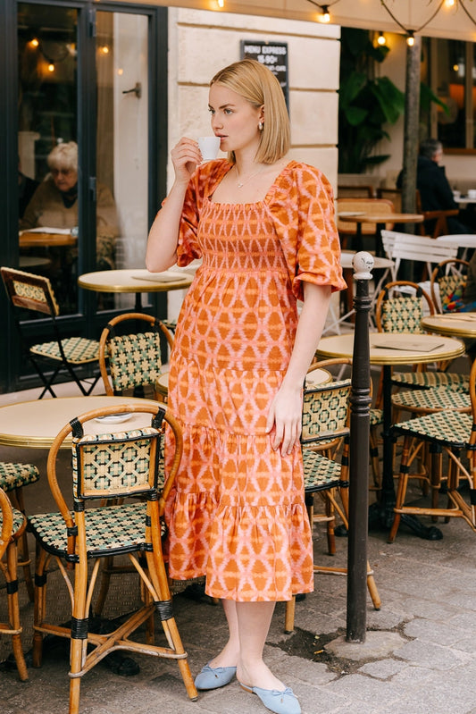 The Smocked Maxi Dress in Munni Sunset is a long dress composed of a rayon-cotton blend fabric. It has a tiered skirt and extends to mid-calf. The fabric features an orange and pink tie-die style geometric print. The dress has a square, elastic lined neckline and loose, elbow-length sleeves.