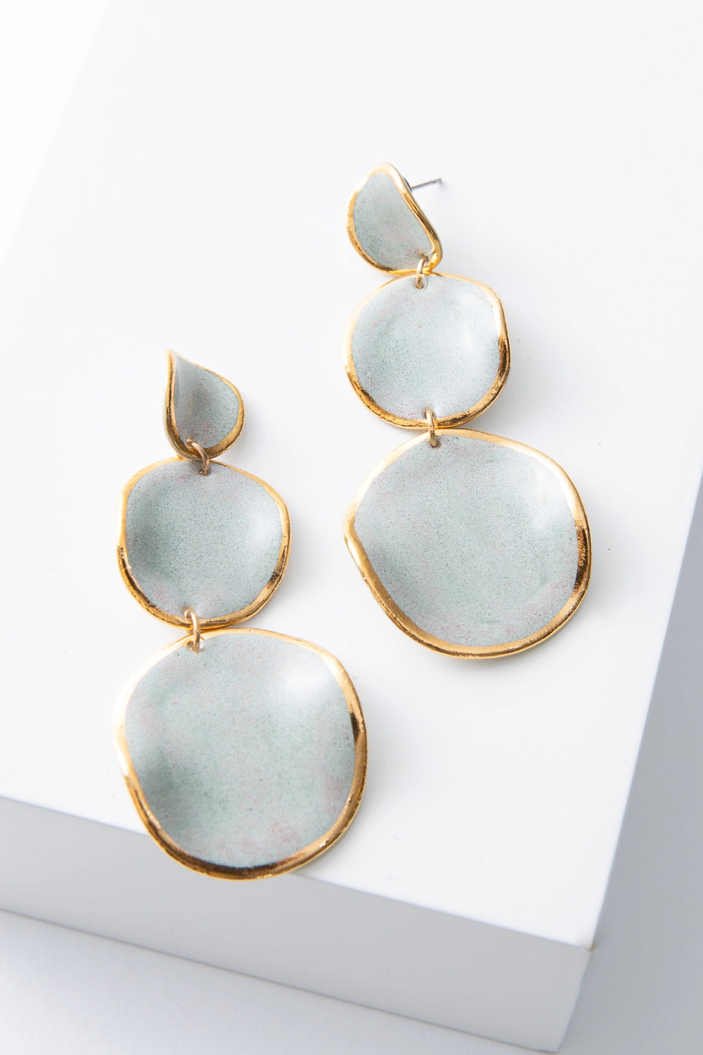 The Stacked Spheres Earrings are dangly earrings composed of three flat, disc-shaped pieces of porcelain stacked on top of each other. The top disc is small, and the discs get larger as you move down the earring. Each disc is composed of light grey porcelain outlined in a ring of shining gold. Rather than being perfectly flat, the discs have a wavy, irregular shape.