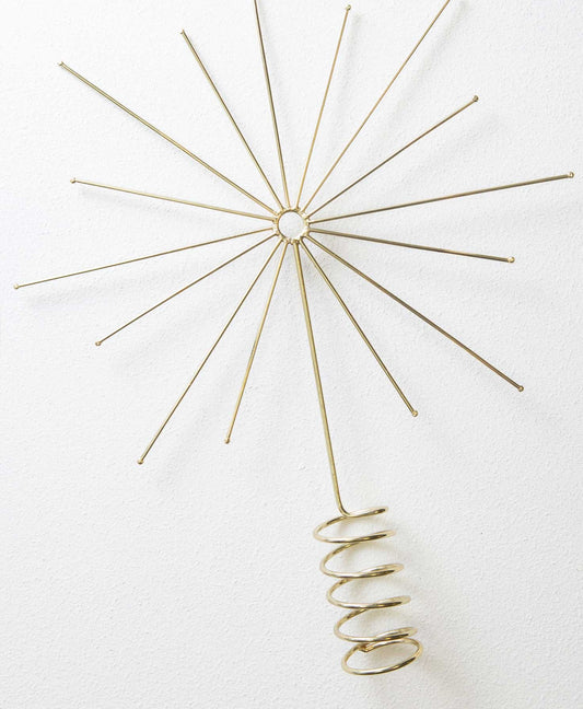 The Starburst Tree Topper is composed of shiny gold metal. It has a coiled base where the topper can be attached to the top of a tree. Above this, the metal forms a minimalist starburst design with a midcentury modern look.