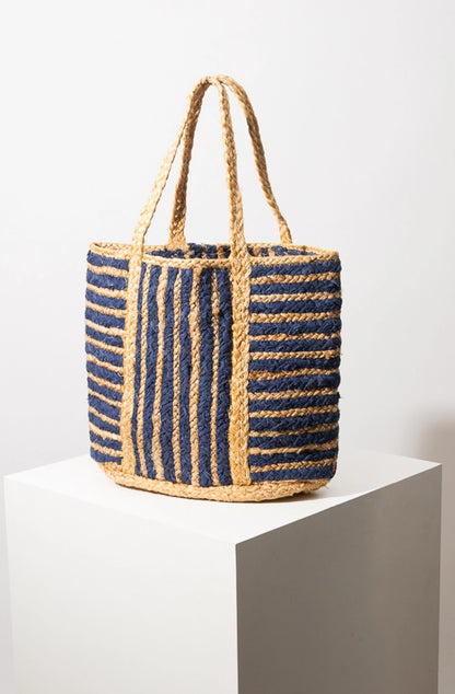 The Striped Jute Tote is a square shaped tote bag composed of braided jute. The structured bag has a natural tan base adorned with horizontal and vertical navy stripes. It has two woven jute handles and is open at the top.