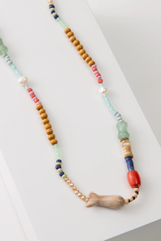 The Surf Side Necklace is a rope necklace composed of sections of many different beads, including paper, wood, fresh water pearl, and glass beads. At the base of the necklace is a large fish bead composed of brown horn. The colors in the necklace include mint, pink, lavender, blue, and red.