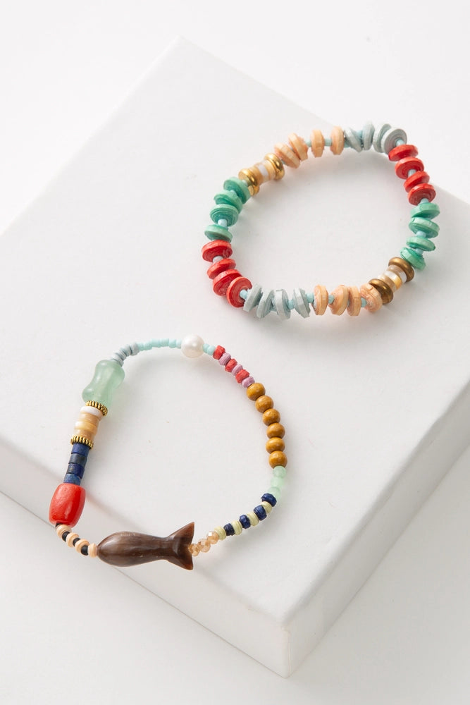 The two bracelets included in the Surf Stack are both single strand bracelets. One is composed of disc-shaped paper beads in shades of tan, light blue, coral, and mint, interspersed with brass and horn beads. The other is composed of many different beads, including paper, wood, fresh water pearl, and glass beads. It also has a large fish bead composed of brown horn. The colors in the bracelet include mint, pink, lavender, and blue.