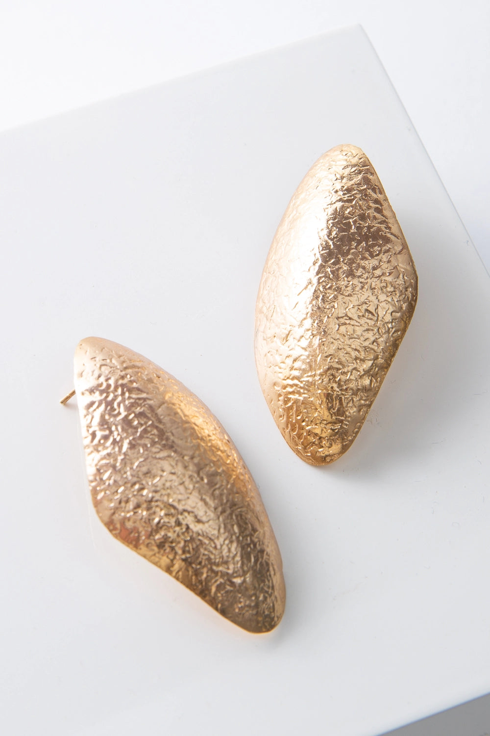These are post-style earrings composed of shining gold-plated brass with a hammered texture. The earrings have an irregular, organic look and are roughly diamond shaped, with smooth rounded edges.