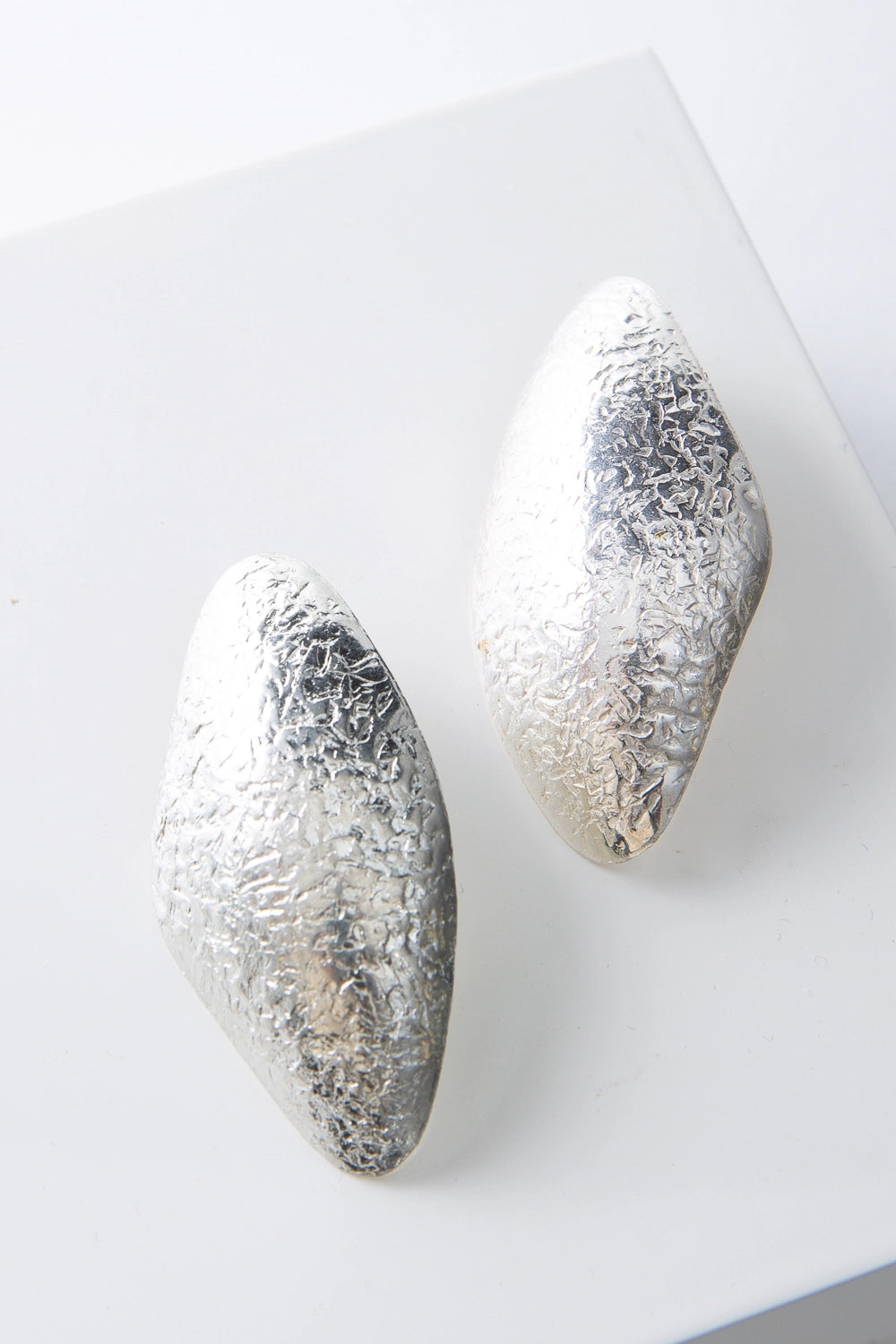 These are post-style earrings composed of shining silver-plated brass with a hammered texture. The earrings have an irregular, organic look and are roughly diamond shaped, with smooth rounded edges.
