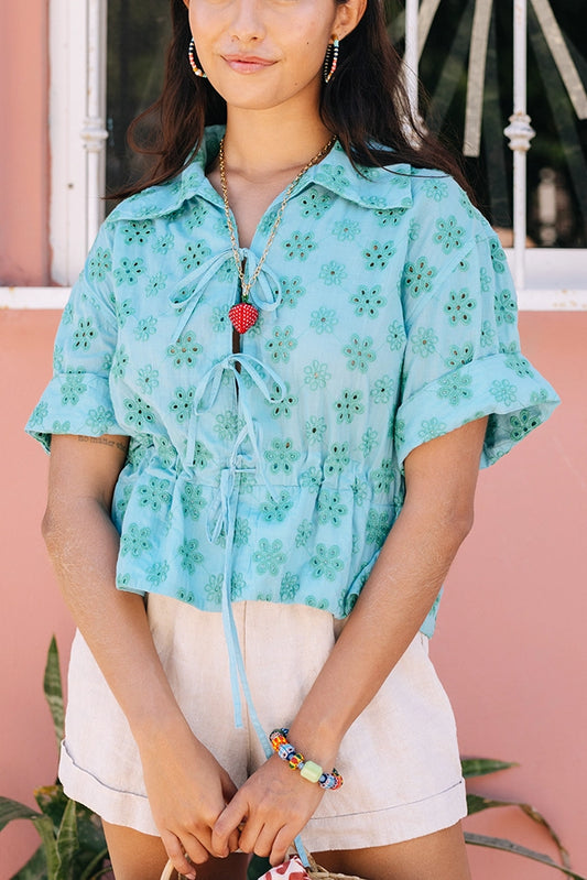 The Tie Cinch Blouse is a short-sleeve top composed of blue cotton fabric covered in a floral eyelet pattern. There is a drawstring at the waist that can be tightened and tied, creating a ruffled look below it. The top has a collar and is open down the middle, with two bow tie closures.