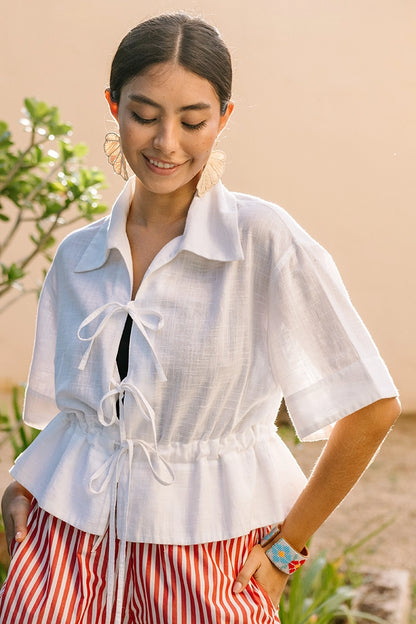 The Tie Cinch Blouse is a short-sleeve top composed of crisp white cotton fabric. There is a drawstring at the waist that can be tightened and tied, creating a ruffled look below it. The top has a collar and is open down the middle, with two bow tie closures.