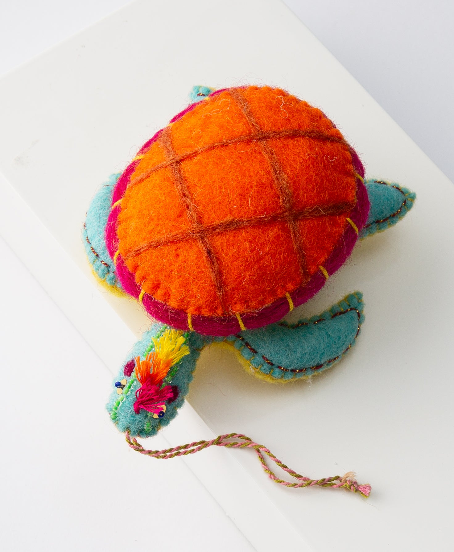 The Tiptop Turtle Ornament is shown from overhead. It is a small stuffed turtle made of fabric. The shell is bright orange and hot pink. The turtle is teal with a yellow belly. Festive thread fringe sticks out from the top of the turtle's head. A braided string attached to the body allows the ornament to be hung.