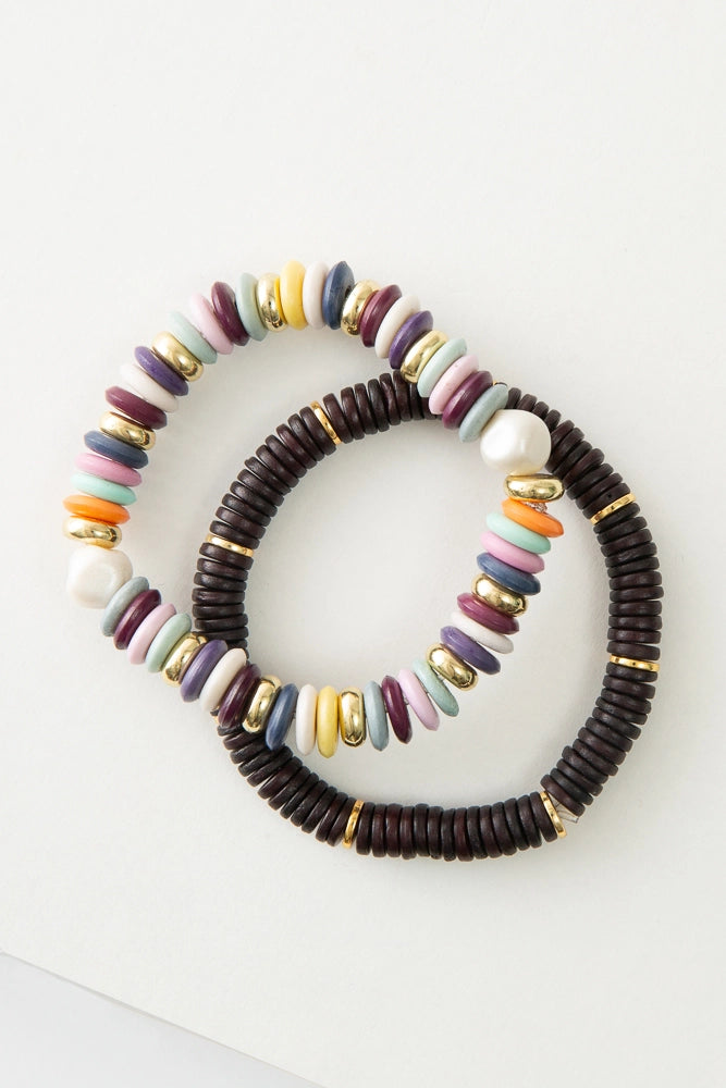 The two bracelets in the Total Eclipse Bracelet Set are each single stranded beaded bracelets. One bracelet features dark brown discs composed of bone, plus brass disc accents. The second bracelet is composed of glass disc beads in shades of orange, pink, mint, purple, maroon, and white, plus brass and pearl accents.